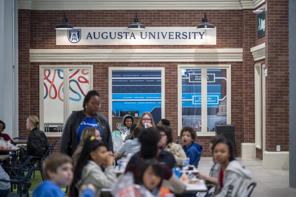 A large room is filled with middle school students eating lunch with a store front in focus in the background that shows a logo for Augusta University.