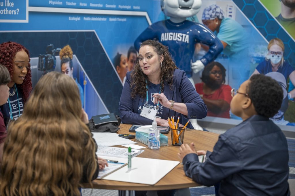 A woman in a blue Augusta University t-shirt and blue blazer sits at a round table with students talking to them. A statue of Augustus with his arms on his hips stands behind her in the corner.