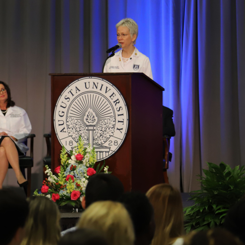 A woman in a health care white coat with a logo for Augusta University stands at a podium and speaks to a large group of people.