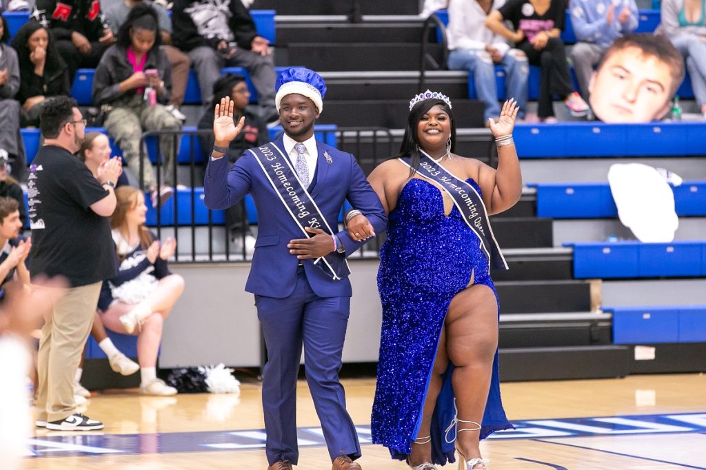 Last year's Homecoming king wearing a blue suit, sash and crown and Homecoming queen wearing a blue sequinned dress, sash and crown wave to the crowd as they walk onto the basketball court