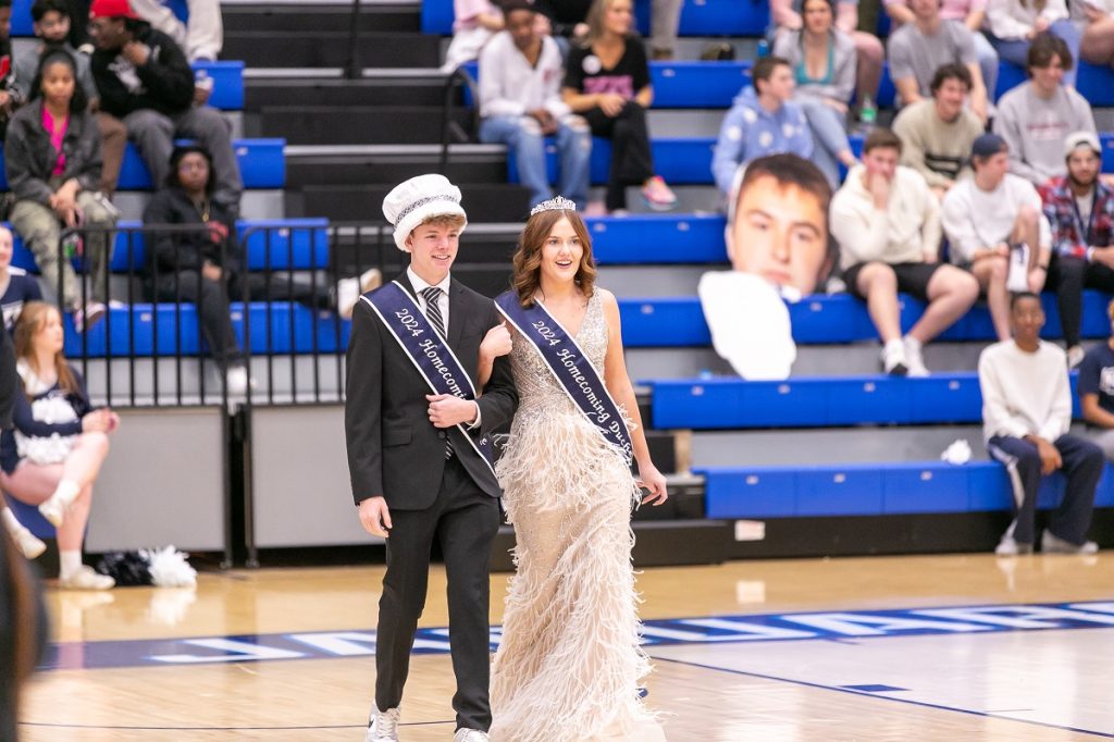 A young man in a black suit, white top-hat crown and sash is arm-in-arm with a young woman in a feathery light colored dress, crown and sash walk onto the basketball court as the crowd looks on