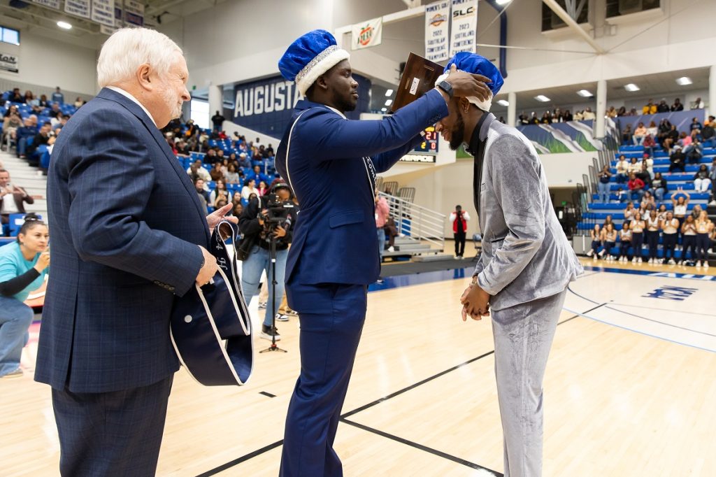 Last year's Homecoming king dressed in a blue suit crowns the new Homecoming king dresses in a silvery gray velour suit while a man in a blue suit stands behind them waiting with a sash in the middle of the basketball court while the spectators in the stands cheer
