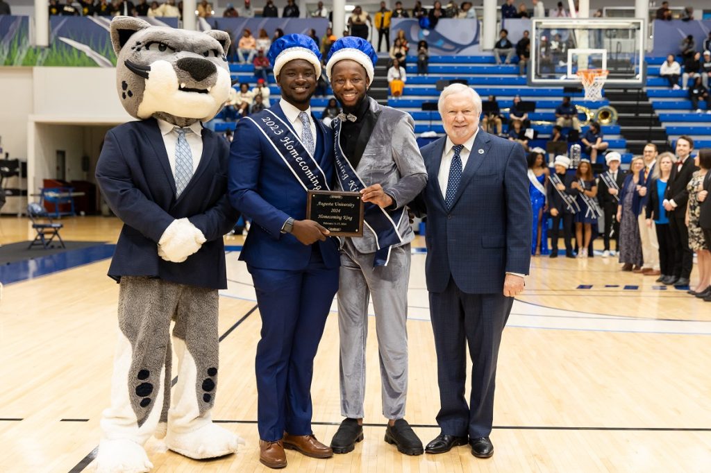 The Augusta University mascot dresses in a suit coat, shirt and tie poses with the past and present Homecoming king dressed in suits with crowns and sashes and holding a Homecoming king plaque and President Brooks A. Keel, PhD, in the middle of the basketball court