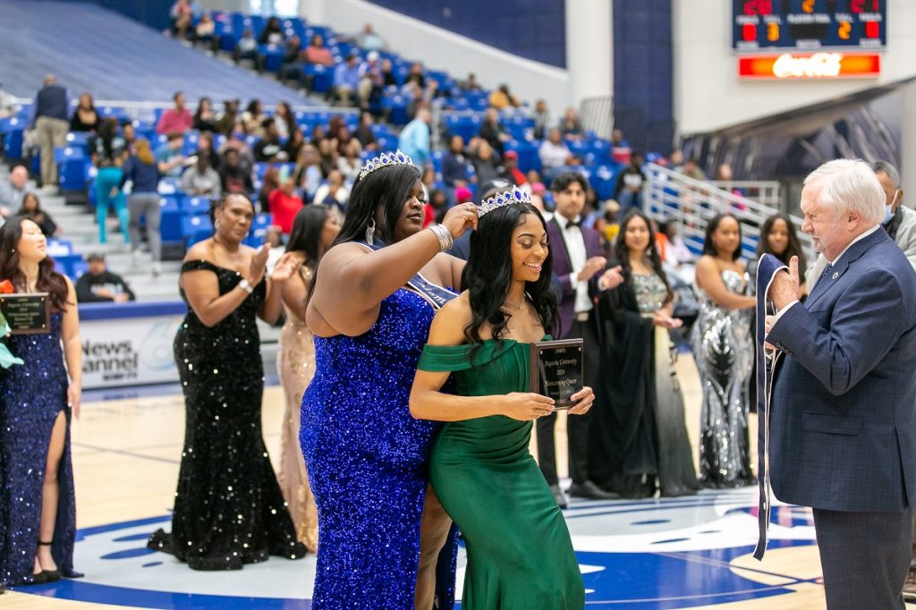 Woman in a royal blue sequinned dress places a crown on the head of a woman in an off-the-shoulder green dress holding a homecoming queen plaque while a man in a dark blue suit stands in front of them with a sash and other women in fancy dresses clap in the middle of the basketball court
