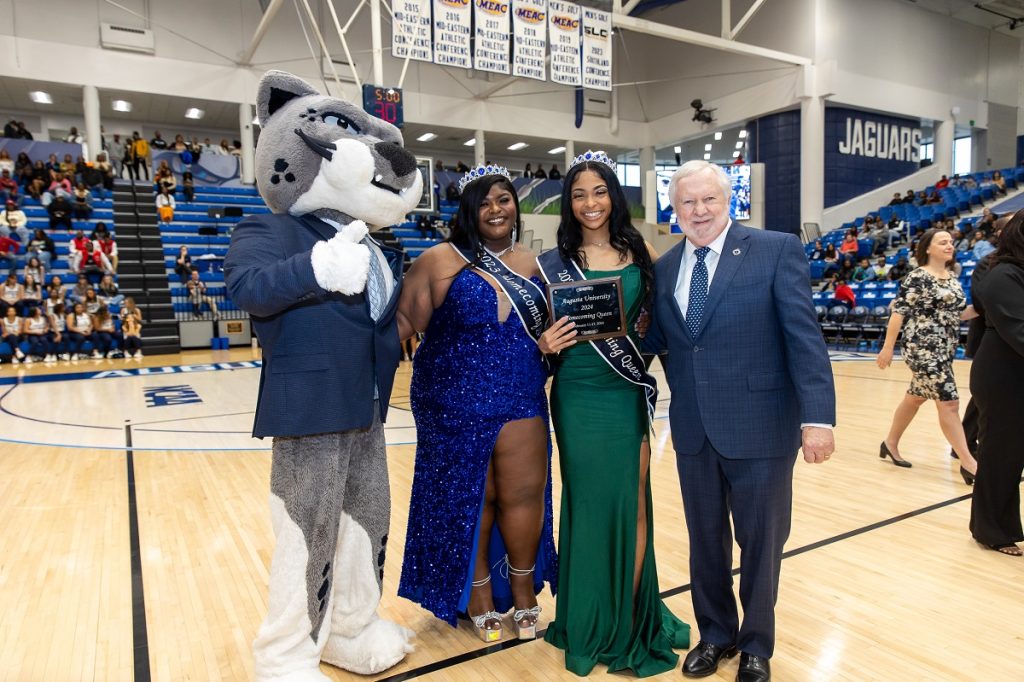 Augustus the mascot dressed in a suit coat, short and tie poses with the past and new homecoming queens wearing dresses, crowns, sashes and holding a homecoming queen plaque and President Brooks A. Keel
