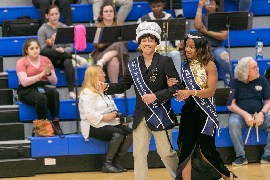 Two college students, a man and a woman, wearing homecoming sashes and crowns walk along the side of a basketball court.