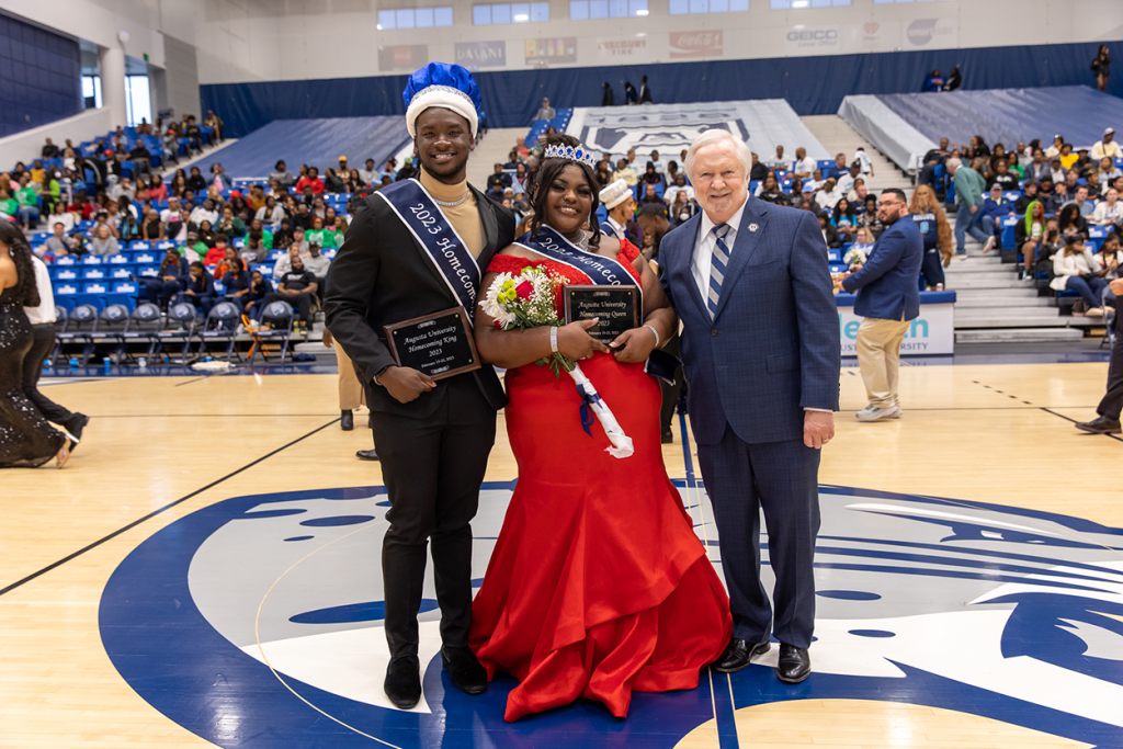 A male college student and a female college student wearing homecoming sashes and crowns pose for a picture with a man in a suit on a basketball court.