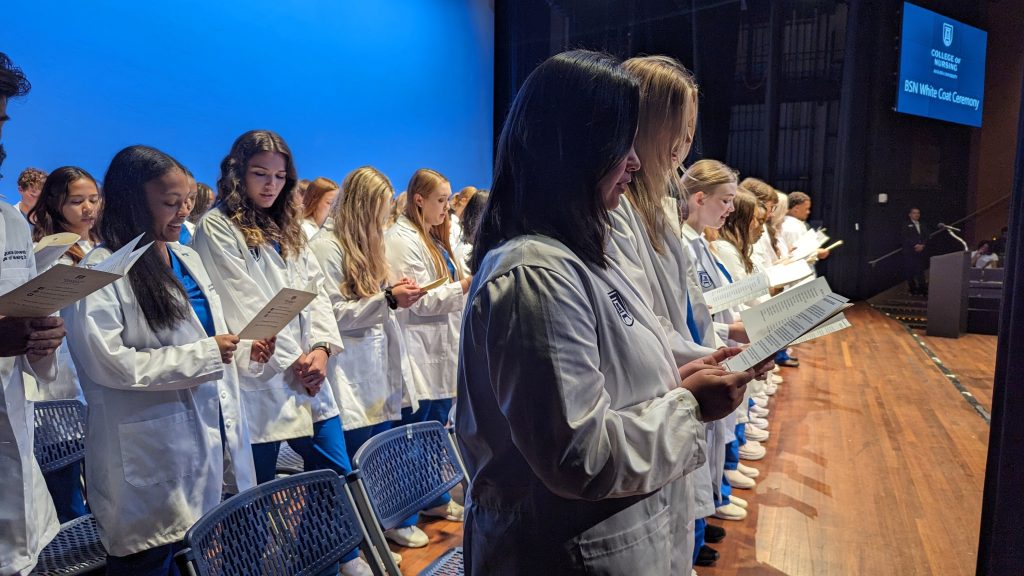 Nursing students stand and recite an oath while on a stage after receiving their clinical coats.