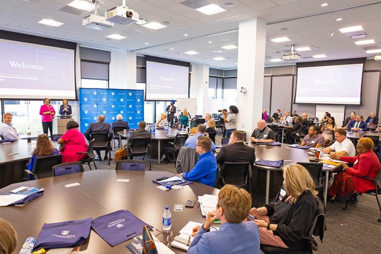 A large conference room is filled with people sitting at tables listening to a presentation.