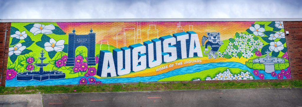 Completed mural with a fountain, white and pink flowers, a navy Augusta University bell tower, the words Augusta Home of the Jaguars with city building outlined above the word Augusta within a pink, orange and yellow sunset, Augustus the mascot, a river flowing at the bottom of the mural, with more flowers and bright green grass throughout
