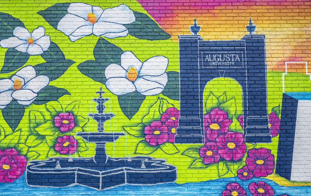A closeup of one of the sections of the mural with white magnolia flowers, a fountain, bright pink flowers in the bright green grass and a navy blue Augusta University arch