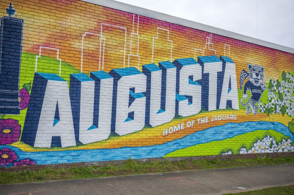 A closeup of part of the mural that says Augusta Home of the Jaguars with Augustus the mascot at the end of the word Augusta. A skyline of buildings against a pink, orange and yellow sunset come out of the top of the word Augusta