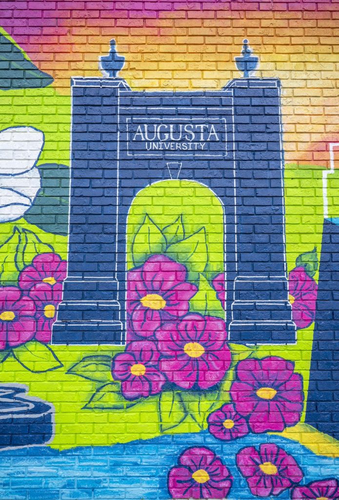 The Augusta University arch from the Summerville Campus is painted in navy blue with white outline and the words Augusta University in white with a neon green background and big bright pink flowers around the bottom of it