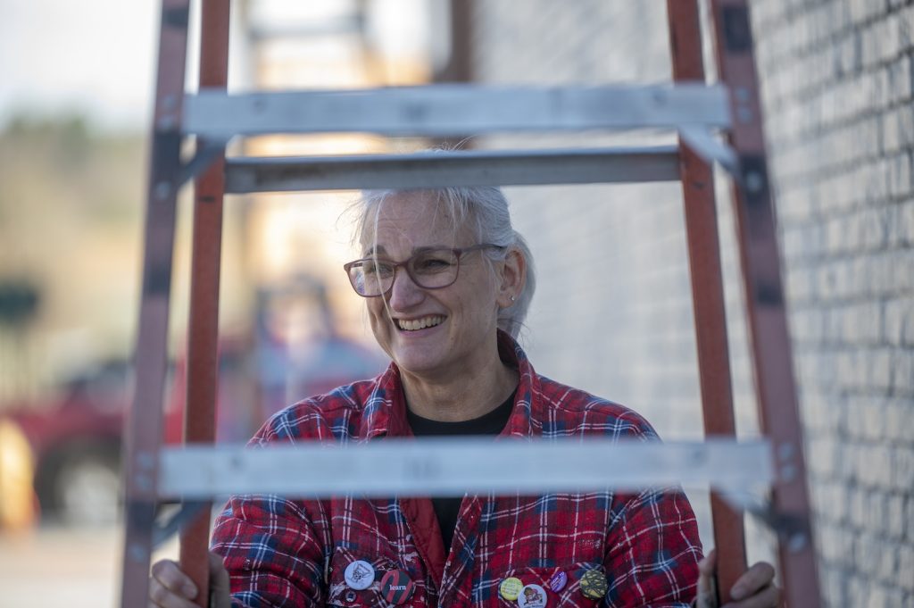 a woman in a red plaid flannel shirt smiles as she moves a ladder in front of the mural