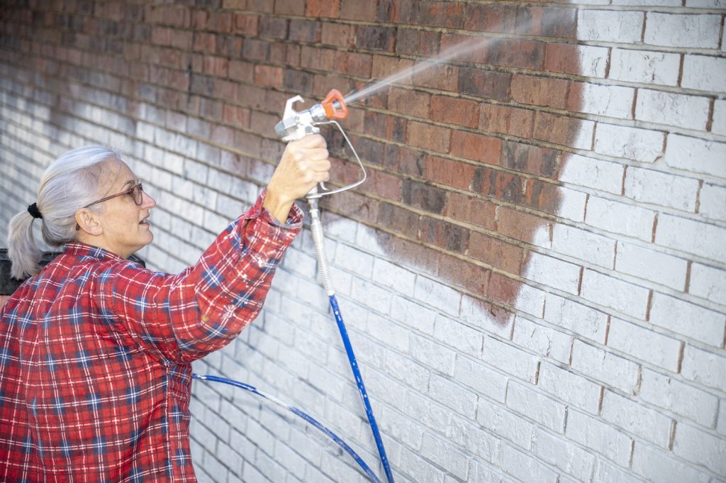 A woman in a red plaid flannel shirt holds a paint sprayer and sprays the brick white