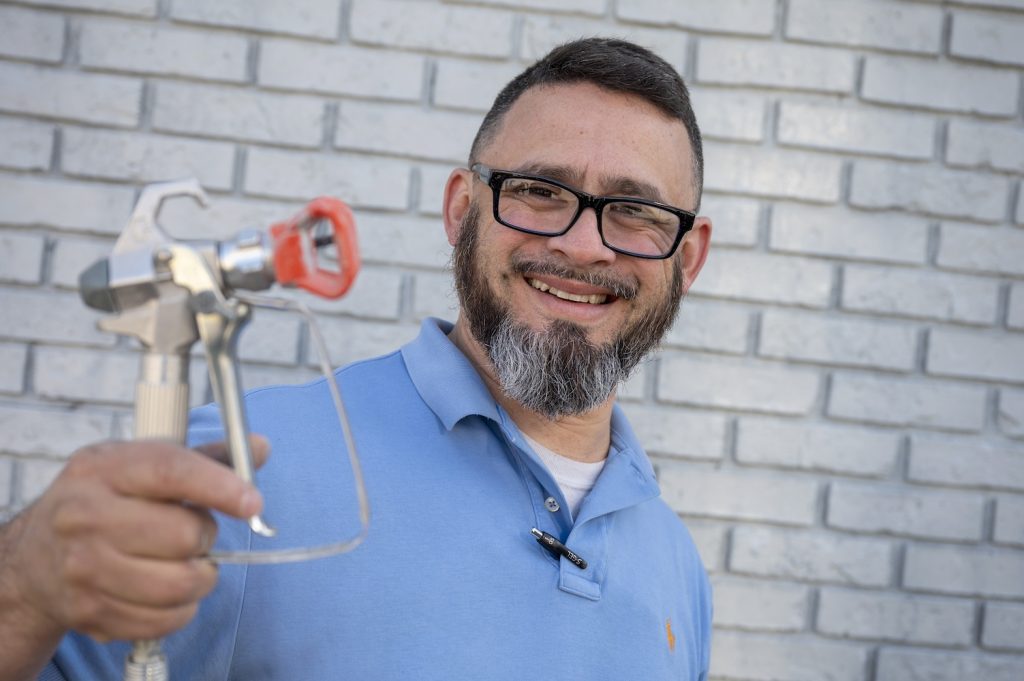 Man in a light blue polo shirt poses with a paint sprayer and smiles at the camera in front of the wall