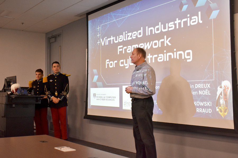 Three men stand at the front of a classroom, two of them in French military regalia, presenting in front of a screen.