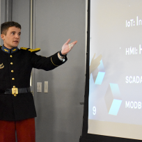 Two men in military uniforms stand at the front of a classroom, presenting in front of a screen.