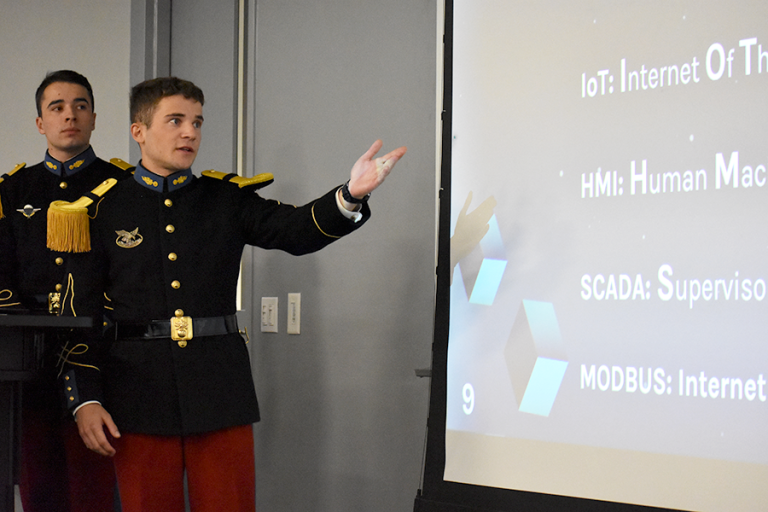 Two men in military uniforms stand at the front of a classroom, presenting in front of a screen.