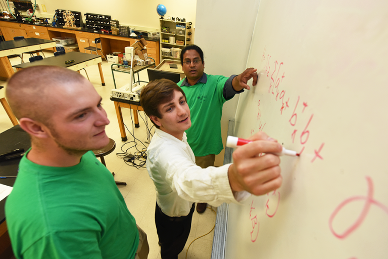 Two college students work with a professor while writing on a white board in a classroom.