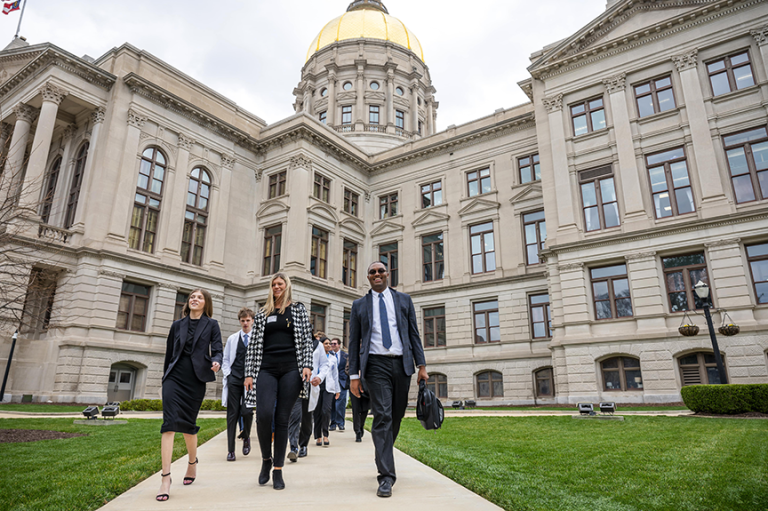 A large group of people walk outside in front of a large state capitol building.