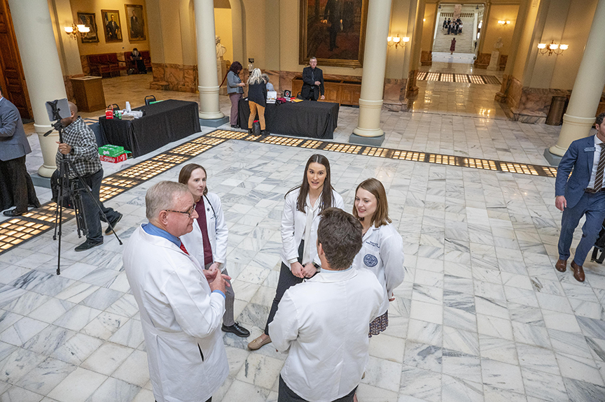 Five people wearing health care professionals coats stand inside a large foyer talking while people set up tables for presentations around them.