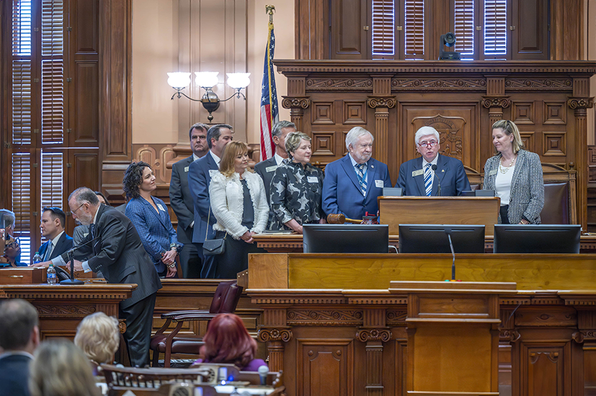 Nine people stand at a large, ornate podium inside a State Capitol, listening to the man on the right in front of the microphone speaking