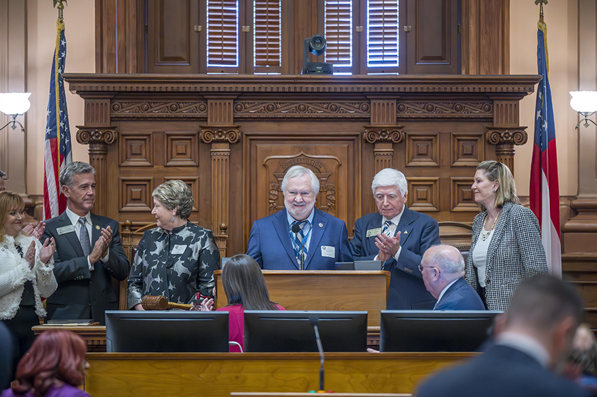 Six people stand at a large, ornate podium inside a State Capitol, applauding the man in the middle, AU President Brooks A. Keel, PhD