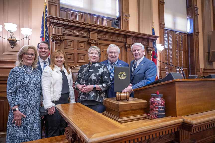 Six people pose for a photo at a large, ornate podium with a gavel and black leather-bound folder with a gold emblem
