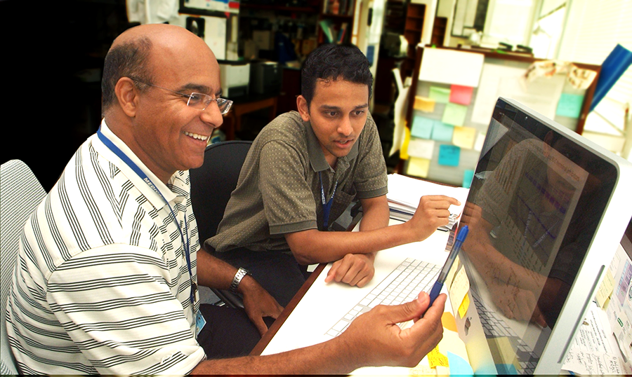 Two men sit at a desk with a computer. One of the men uses a pen to point at the computer's screen showing data graphs.