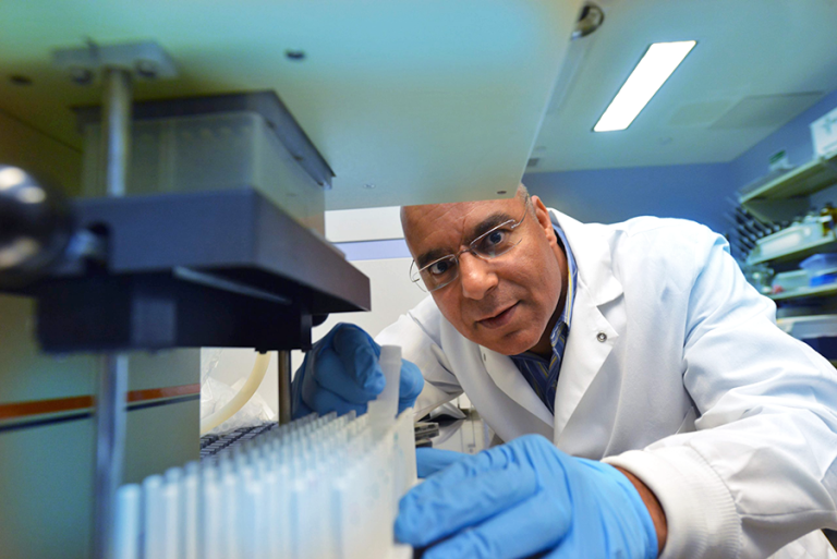 A male medical researcher wearing a white medical coat and blue gloves looks into a machine with lots of plastic tubes.