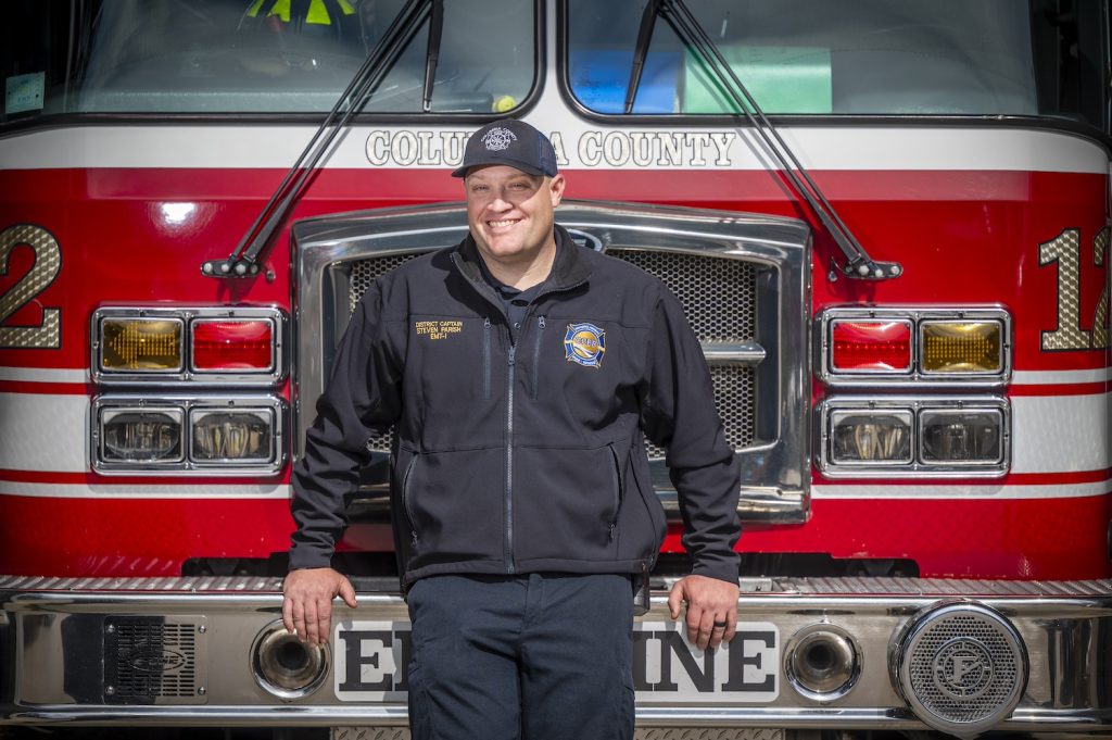 Captain in his department-issued jacket and hat leans against the front of a fire truck smiling at the camera.