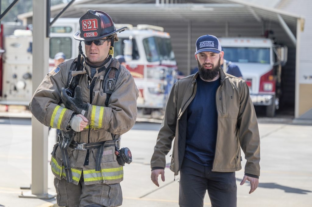 a man in full firefighter attire walks next to a man in a navy blue shirt, tan jacet and blue Augusta University hat