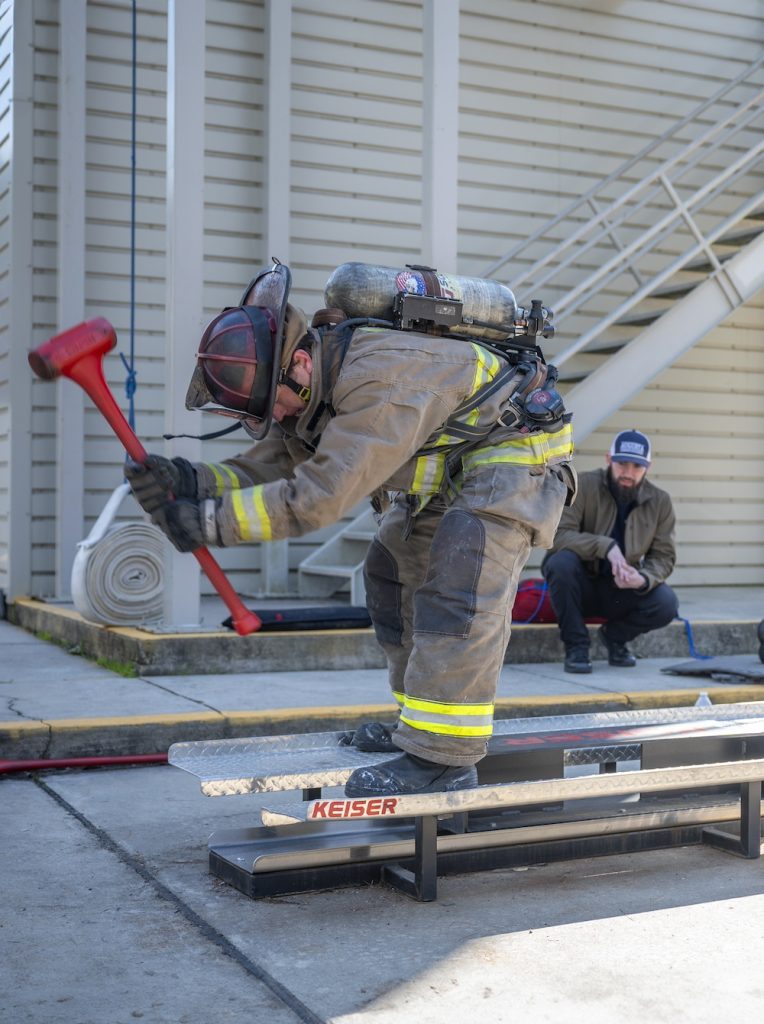 Firefighter in full gear bends over while holding a red mallet standing on a metal platform with the word Keiser in red while a man in an Augusta University hat crouches down behind him watching