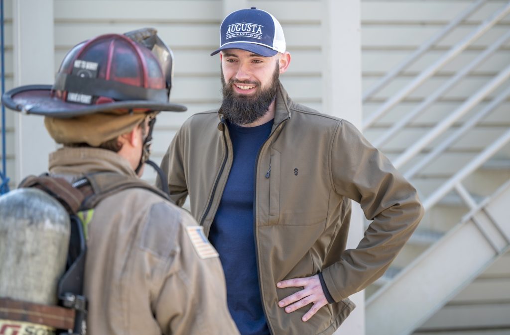a man in a navy blue t-shirt, tan jacket and blue and white Augusta University hat stands smiling with his hands on his hips talking to a firefighter in full gear