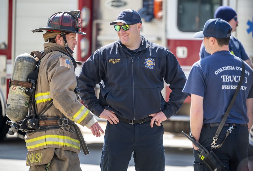 A man in the center with his department-issued jacket, sunglasses and a hat smiles as he talks to a firefighter in full gear to the left and another man in a blue Columbia County Truck 1 Fire Rescue and hat faces away from the camera toward them listening to the conversation