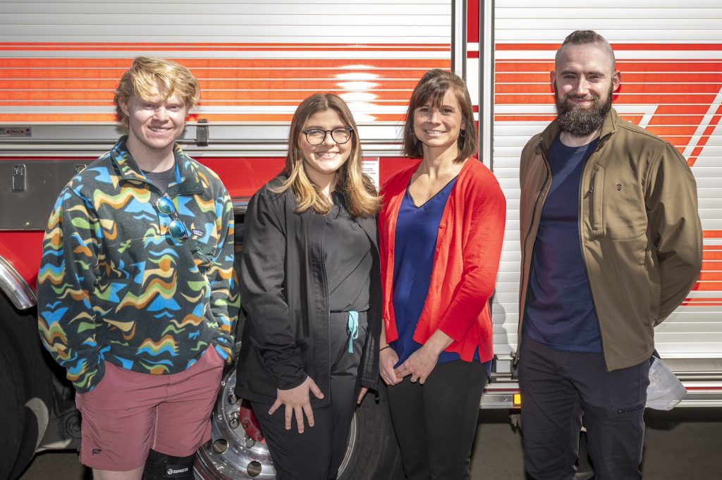 a man in a black sweatshirt with blue, orange, yellow and green designs, a woman in black scrubs and a black jacket, a woman in a navy blue shirt and bright red sweater and a man in a navy blue t-shirt and tan jacket stand next to a fire truck smiling at the camera