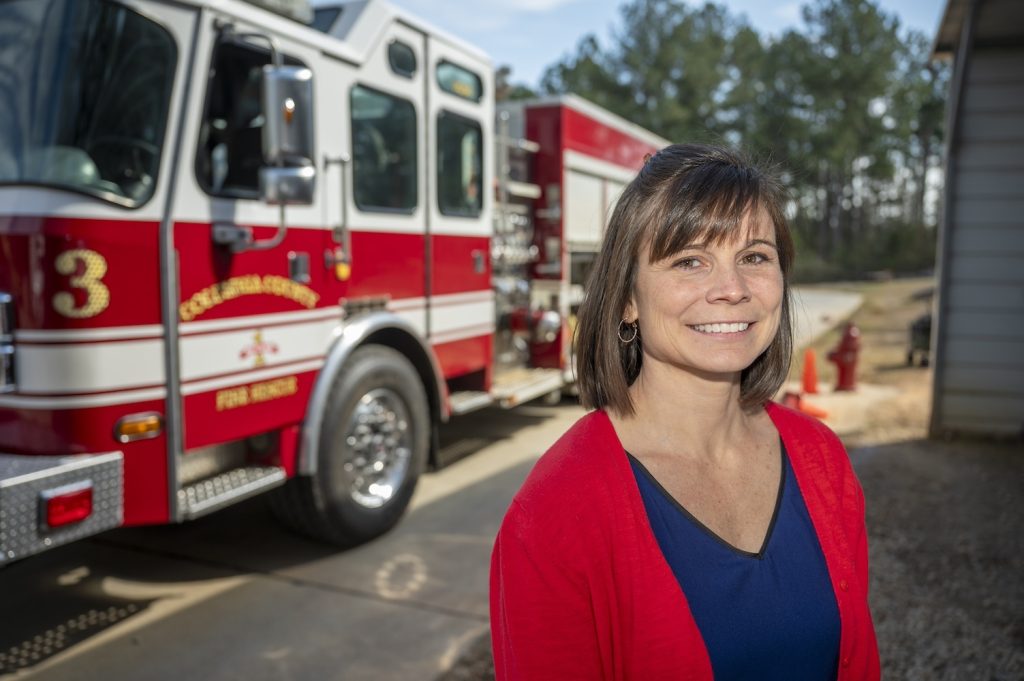 Woman in a navy blue shirt and bright red sweater stands in front of a fire truck smiling at the camera