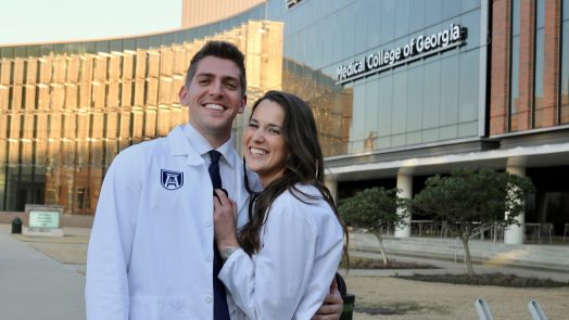 A young couple in white coats pose in front of the Medical College of Georgia