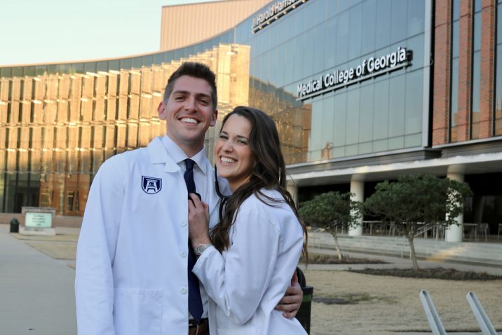 A young couple in white coats pose in front of the Medical College of Georgia