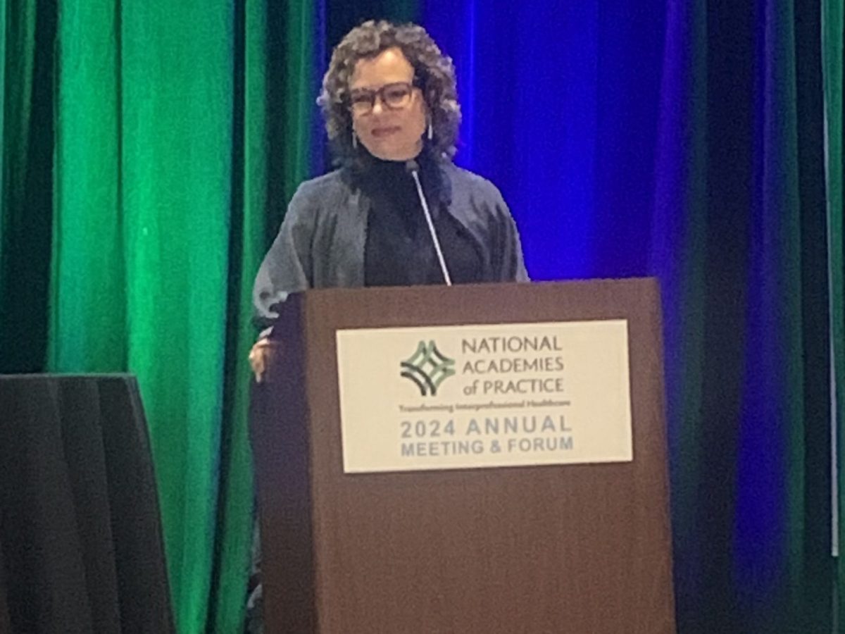 a woman stands behind a podium speaking with a green and blue curtain behind her. On the podium is a gold plaque that reads "National Academies of Practice -- transforming interprofessional healthcare -- 2024 annual meeting & forum"