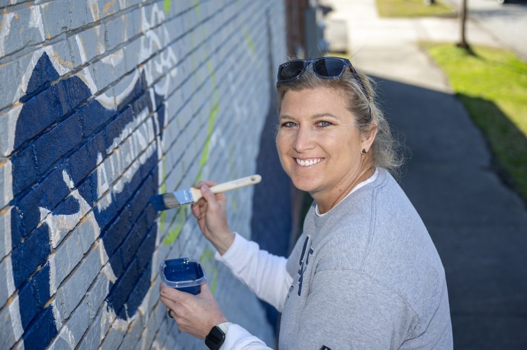 Woman in a gray Augusta University t-shirt smiles at the camera while painting Augusta University's mascot Augustus in the mural