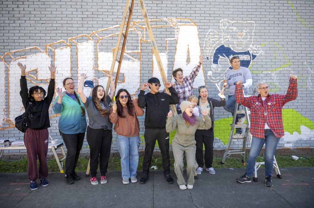 Nine women, one male and a toddler cheer with unfinished mural in progress behind them