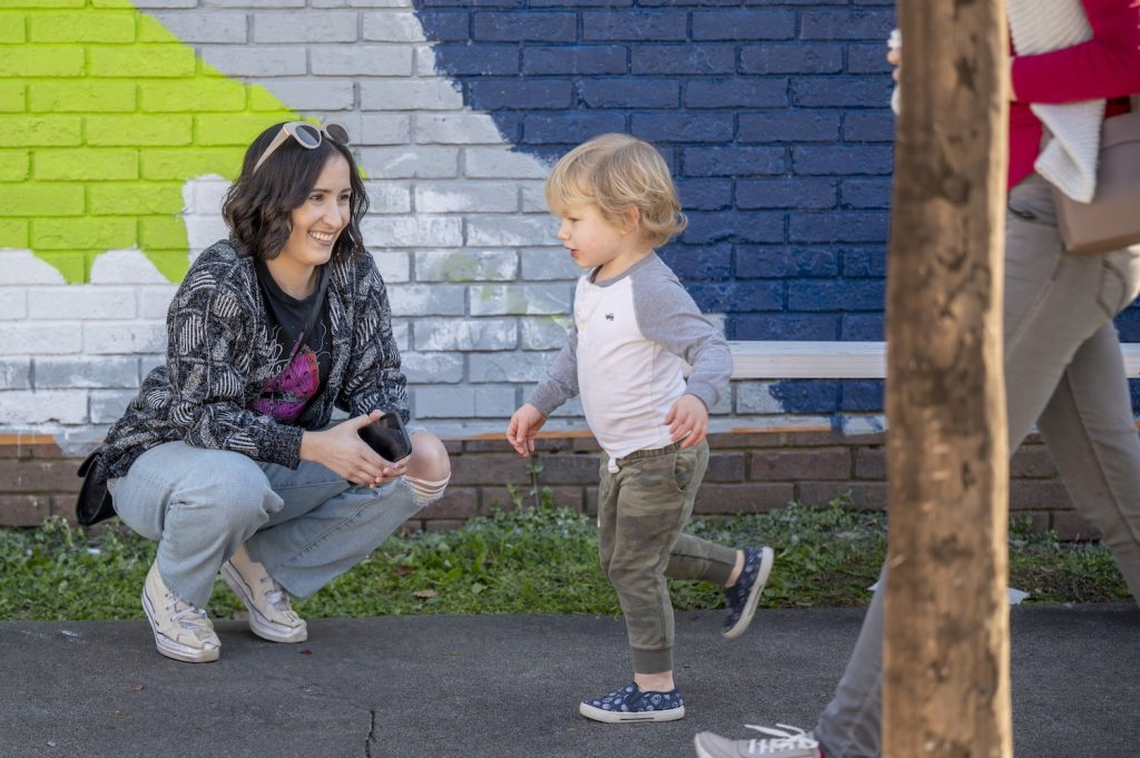 Woman crouched down smiles as a child runs by in front of the mural