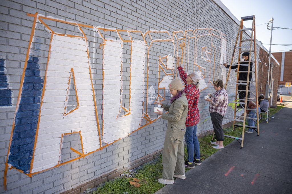 Three women on the ground paint various letters in the word Augusta white while a fourth person on a ladder paints the last A