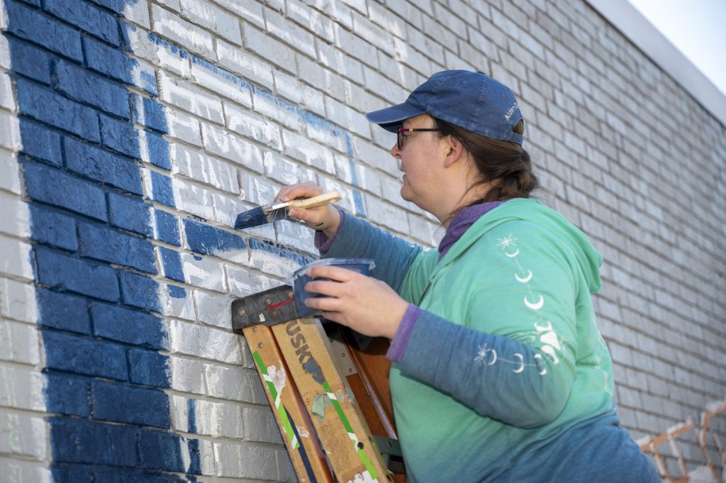 A woman in a teal and blue shirt stands at the top of a ladder painting a navy blue bell tower in the mural