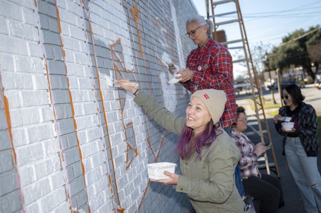 A woman in a tan beanie and olive green jacket smiles while painting the G in Augusta white while another woman in a red plaid flannel shirt paints the S in Augusta white behind her