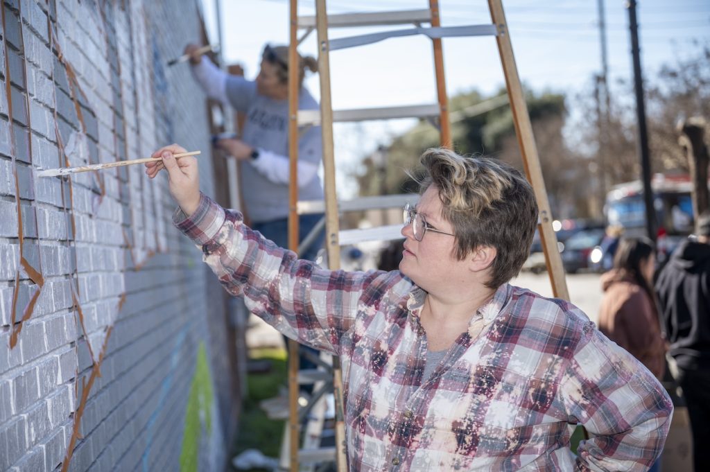 A woman with a flannel shirt covered in paint paints the second letter U in the word Augusta on the mural