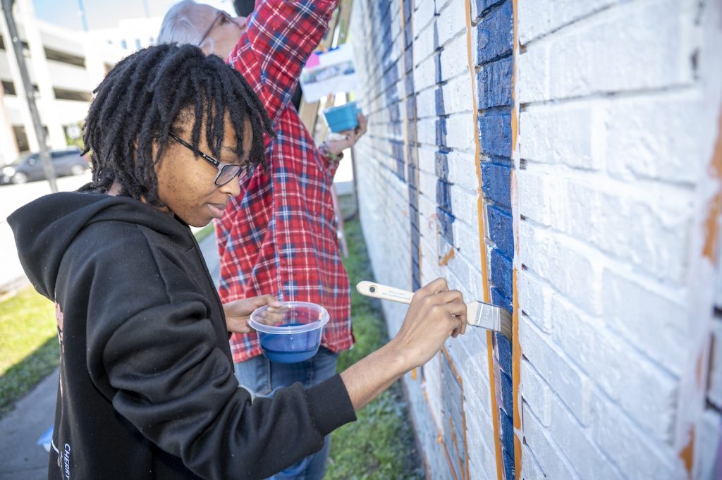 A woman in a black hoodie paints blue between the white letters of the word Augusta on the mural