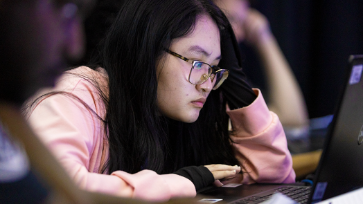 A female college student rests her head on one of her hands as she concentrates on reading a computer screen.